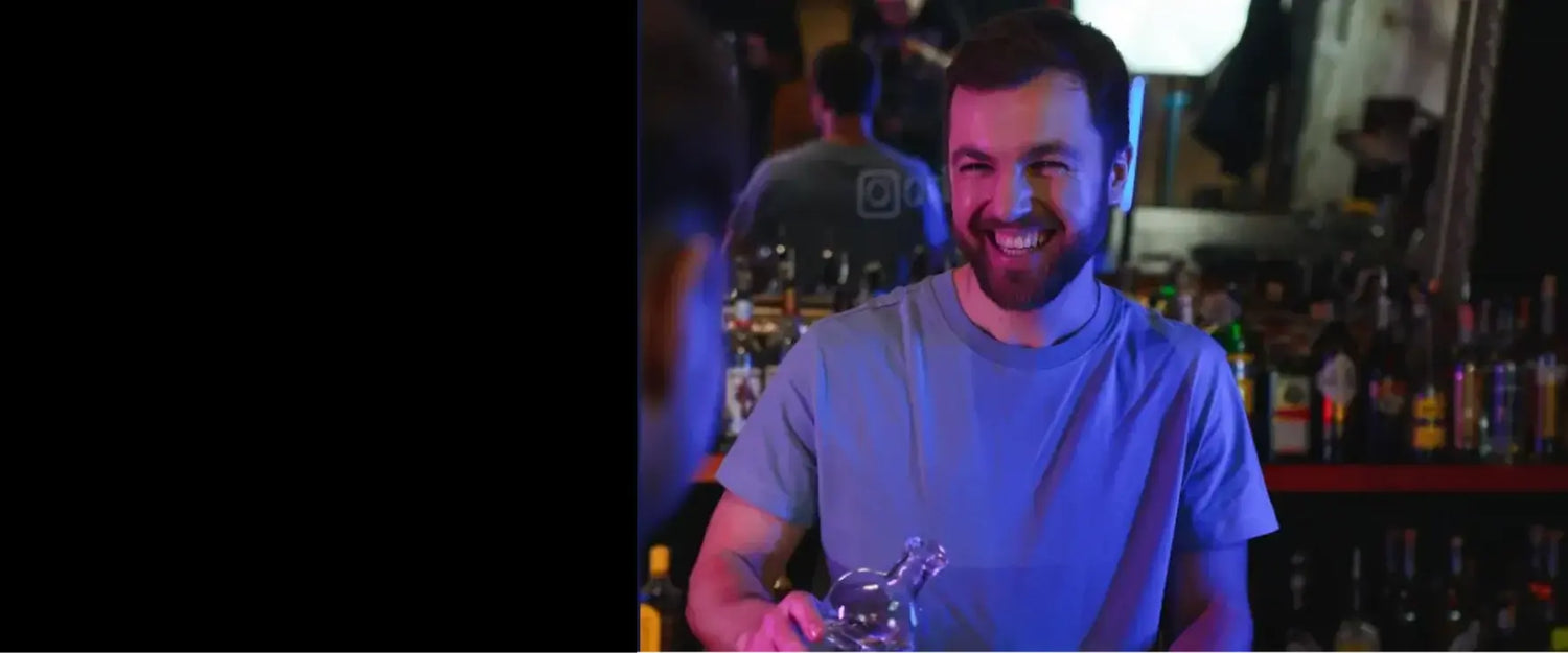A man with a beard and a blue shirt, standing laughing in a bar setting